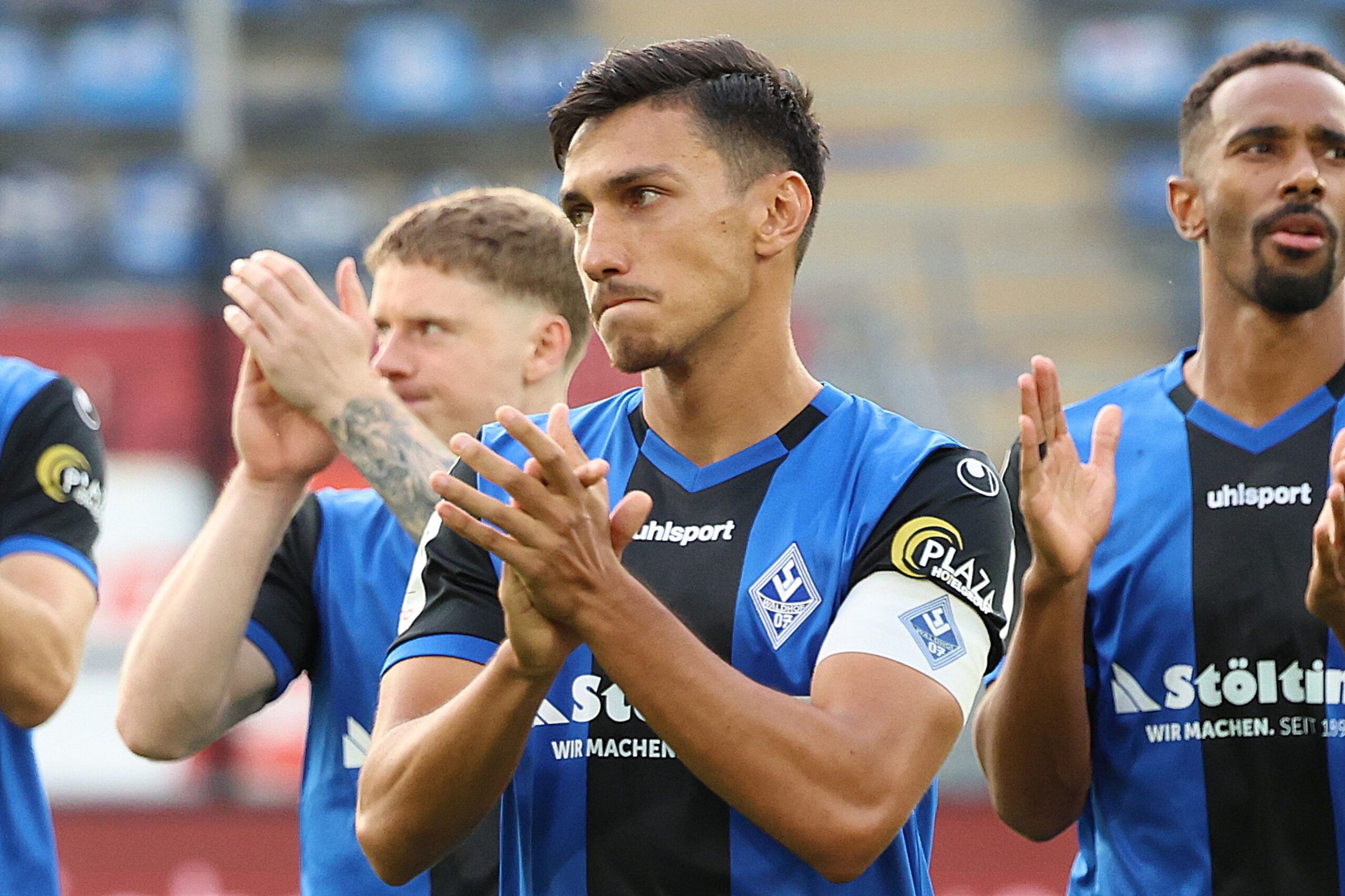 Mehrere Fußballspieler des SV Waldhof Mannheim in blauen Trikots mit schwarzen Streifen applaudieren stehend auf dem Spielfeld.