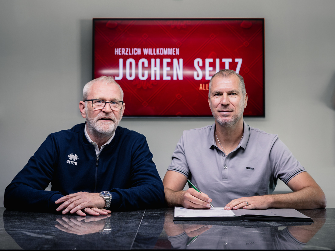 Zwei Männer sitzen an einem Tisch. Der Mann rechts trägt ein helles Poloshirt und unterschreibt ein Dokument, der Mann links trägt eine dunkle Jacke und eine Brille. Im Hintergrund ist ein roter Bildschirm mit der Aufschrift »Herzlich Willkommen Jochen Seitz« zu sehen.